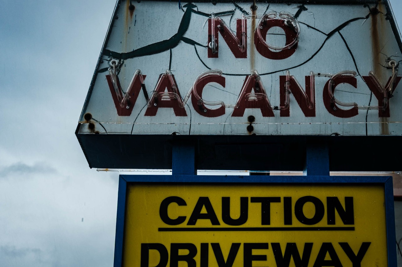 A weathered roadside sign reading ‘No Vacancy’ above ‘Caution Driveway.’